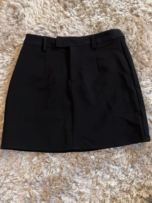 Svart ”kostymkjol” från Bik Bok - Stilren svart kortkjol med bälteshällor och diskreta fickdetaljer. Kjolen har en rak passform och är perfekt att matcha med olika toppar. Materialet känns mjukt och har en lätt stretch för extra komfort.