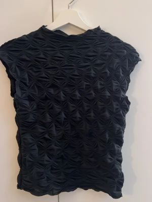 Svart topp med struktur från Gina Tricot - Säljer en svart topp från Gina Tricot i storlek S. Toppen har ett coolt strukturerat tyg med 3D-effekt och korta ärmar. Materialet är stretchigt och ger en unik look som sticker ut. Perfekt snygg svart topp!