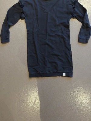 Svart/grå klänning från I Dig Denim - BARN: En enkel och stilren mörkblå klänning från I Dig Denim med långa ärmar och diskreta volangdetaljer på axlarna. Tillverkad i mjuk bomull, perfekt för en avslappnad och snygg look.