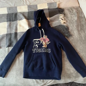 Marinblå hoodie Polo Ralph Lauren - Snygg marinblå hoodie från Polo Ralph Lauren med tryck av en tiger och texten 'TIGERS' på bröstet. Ficka framtill och justerbar huva med vita snören. Väldigt tjockt och skönt material.