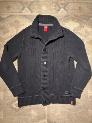 Mörkgrå kabelstickad kofta från Esprit - Snygg mörkgrå kabelstickad kofta från Esprit med stora knappar och bred ribbad krage. Koftan har långa ärmar, ribbade muddar och detaljerad stickning framtill. Perfekt för lager på lager och ger en stilren look.