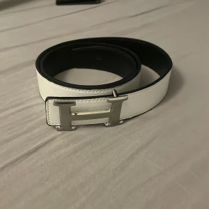  vitt skärp med silver H-spänne - Snyggt och stilrent skärp från H i vitt skinn med ett stort H-format spänne i silver. Bältet är vändbart med svart insida och har klassisk design som passar till många outfits. Perfekt accessoar för dig som gillar exklusiva detaljer.