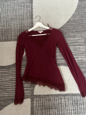 Vinröd långärmad topp Gina Tricot - Supersnygg vinröd långärmad topp från Gina Tricot med spetsdetaljer vid halsringning och nederkant. Toppen har en v-ringad form och är tillverkad i ett mjukt, stretchigt material som sitter tight och bekvämt.