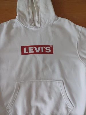 Vit hoodie från Levi's strl M - Säljer en vit hoodie från Levi's med den klassiska röda loggan tryckt på bröstet. Hoodien har huva och en stor magficka framtill. Perfekt för dig som gillar en clean och stilren look med streetkänsla.