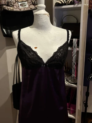 Cubus Satin Nattlinne / Satin Slip Dress - Elegant satin nattlinne/slip dress från Cubus i en djup mörklila färg med vackra spetsdetaljer vid bysten. Det mjuka satinliknande tyget ger plagget en feminin och lyxig känsla. Designen har tunna axelband och en dekorativ rosett baktill som ger en fin detalj. Perfekt som nattlinne, loungewear eller som en romantisk slip dress.