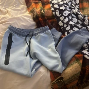 Ljusblå Nike tech byxor från Nike  - Snygga ljusblå nike tech byxor  med svart snörning i midjan och en lång svart dragkedja på ena benet. Byxorna har ribbade muddar vid bensluten och klassisk Nike-logga tryckt på låret. Är i storlek L men passar även M