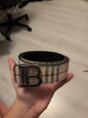 Rutig Burberry bälte med B-spänne - Snyggt bälte från Burberry med klassiskt rutigt mönster i beige, svart och rött. Bältet har ett stort metallspänne i form av bokstaven B och insidan är svart. Perfekt accessoar för att lyfta din outfit.