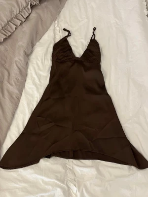 Brun kort klänning med smala band - Snygg brun kort klänning med smala axelband och v-ringad hals. Klänningen har en lätt utsvängd form och tunna band som knyts i ryggen. Materialet ser ut att vara mjukt och har en lätt glans, vilket ger en elegant känsla.