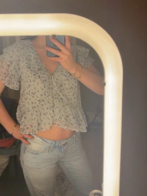 Blommig volangblus från H&M - Söt blus i ljusblått med små mörkblå blommor. Modellen har korta volangärmar, djup v-ringning. Tillverkad i ett tunt, luftigt material som ger en romantisk vibe. Perfekt till jeans eller kjol för en trendig look.