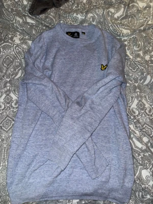 Ljusblå stickad tröja Lyle & Scott - Ljusblå stickad tröja från Lyle & Scott med rund halsringning och klassisk gul logga på bröstet. Tröjan har långa ärmar och ribbade muddar vid ärmslut och nederkant. Perfekt för lager på lager och enkel att matcha.