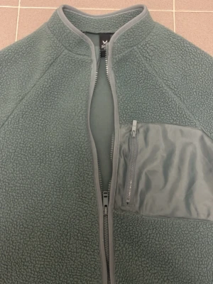 Grön fleecejacka med dragkedja - Mysig grön fleecejacka från SOC med hel dragkedja framtill, bröstficka med dragkedja och sidoficka. Jackan har ribbade detaljer vid krage och ärmslut samt en mjuk, fluffig struktur. Perfekt för lager-på-lager och chill dagar.