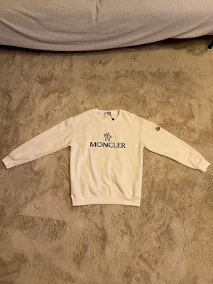 Vit sweatshirt från Moncler - Snygg vit sweatshirt från Moncler med svart logotyptryck på bröstet och Moncler-märke på ärmen. Tröjan har rund halsringning och långa ärmar. Perfekt för en clean och stilren look.