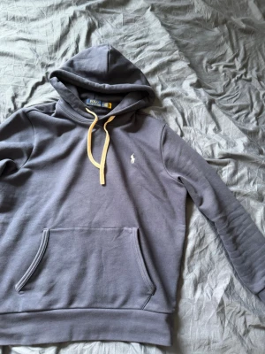 Grå hoodie från Polo Ralph Lauren - Snygg grå hoodie från Polo Ralph Lauren med klassisk huva och beige dragsko. Tröjan har en känguruficka framtill och den ikoniska vita Polo-loggan broderad på bröstet. Tillverkad i mjukt och bekvämt material, perfekt för en avslappnad stil.