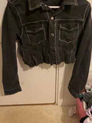 Svart croppad jeansjacka - Säljer en svart croppad jeansjacka med rå kant nertill och vita kontrastsömmar. Jackan har två bröstfickor med lock och knappstängning framtill. Dock har jag klippt den så att den är kortare men de är inget man märker när man har på sig den. Perfekt för dig som gillar en edgy och trendig stil.