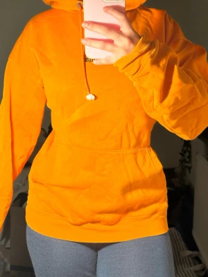 BlACK SQUAD - Snygg orange hoodie med huva och dragsko, samt stor ficka framtill. På ryggen finns ett stort grafiskt tryck i grått och rött med textdetaljer. Hoodien har ribbade muddar vid ärmar och midja. Perfekt för dig som vill sticka ut med färg och cool design.