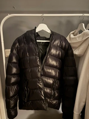 Svart pufferjacka med dragkedja - Snygg svart pufferjacka med glansig finish och raka kanaler. Jackan har två framfickor med dragkedja och en hög krage utan luva. Perfekt för kalla dagar och ger en modern look till din outfit.