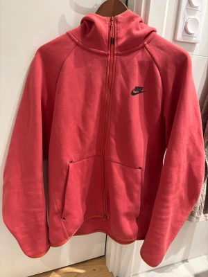 Röd zip hoodie från Nike - Säljer en röd hoodie från Nike med dragkedja framtill och svart Nike-logga på bröstet. Hoodien har huva och två fickor framtill. Tillverkad i mjukt bomullsmaterial som är skönt att ha på sig. Perfekt för en avslappnad och sportig stil.