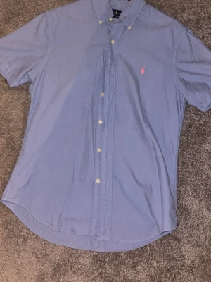 Blå kortärmad skjorta från Ralph Lauren - Snygg blå skjorta från Ralph Lauren med korta ärmar och klassisk button-down krage. Skjortan har knappar framtill och en rosa broderad logga på bröstet. Perfekt för en stilren look.