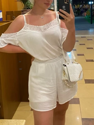 Vit off shoulder jumpsuit med spets - Säljer en vit jumpsuit med off shoulder-design och smala axelband. Har dessutom inbyggda inner-shorts. Jumpsuiten har fina spetsdetaljer vid halsringning och midja samt korta ben. Tillverkad i ett lätt, luftigt tyg som passar perfekt till sommaren. Enkel att styla med accessoarer för en trendig look.
