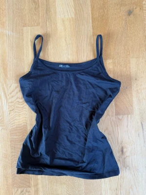 Svart linnetopp från H&M - En enkel och stilren svart linnetopp från Zara med smala axelband. Toppen är figurnära och tillverkad i mjuk bomull, perfekt att matcha med jeans eller kjol. Passar dig som gillar minimalistisk stil.