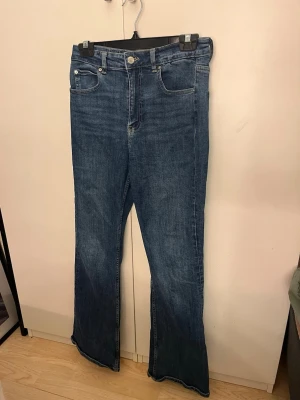 Blå bootcut jeans med hög midja - Säljer ett par klassiska blå bootcut jeans med hög midja och fem fickor. Jeansen har en snygg tvättad look och är tillverkade i denim med stretch. Perfekta för dig som gillar en retroinspirerad stil och vill ha ett par bekväma jeans med lite vidare ben.