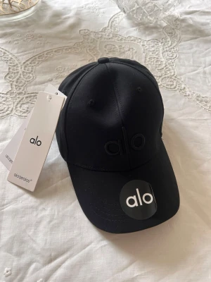 Svart keps från Alo med broderad logga - Snygg svart keps från Alo med broderad logga framtill i samma färg som kepsen. Klassisk böjd skärm och justerbar baksida. Tillverkad i ett mjukt och slitstarkt material som ger en clean och modern look. Perfekt accessoar för en sportig och trendig stil.