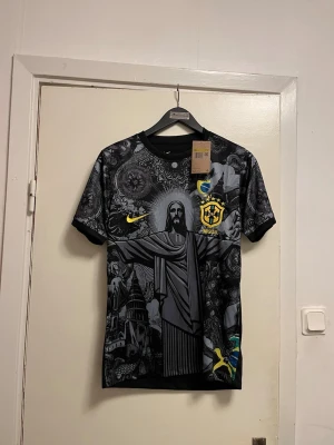 Nike Brazil t-shirt med tryck - Snygg svart t-shirt från Nike med Brasil-tryck och ikoniska motiv som Kristusstatyn och brasilianska symboler i grått och gult. Rund halsringning och korta ärmar. Perfekt för dig som gillar unik design och fotbollsinspirerat mode. Finns i storlek S-M-L