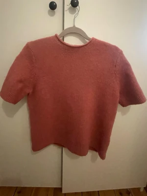 Rosa stickad t-shirt med kort ärm - Mysig stickad t-shirt i en dämpad rosa färg. Tröjan har korta ärmar, rund halsringning och är gjord i ett mjukt, fluffigt material. Perfekt för lager-på-lager och enkel att styla till olika outfits.