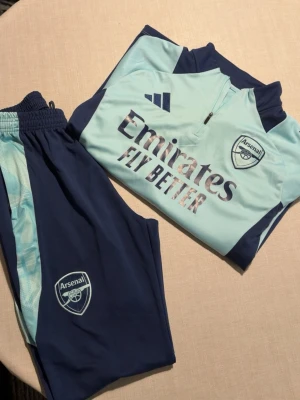 Arsenal träningsset från Adidas - Snyggt Arsenal träningsset från Adidas i ljusblått och marinblått. Setet består av långärmad tröja med halv dragkedja och matchande byxor, båda med Arsenal-logga och Adidas-detaljer. Materialet är funktionspolyester med AEROREADY-teknologi för bästa komfort.