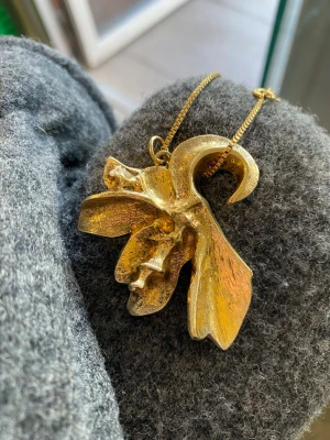 Guldigt halsband med stor blomma - Unikt halsband med ett stort, guldigt blomformat hänge i metall. Hänget har en abstrakt och konstnärlig design med mjuka, böljande former som påminner om en blomma eller orkidé. Kedjan är tunn och matchar hängets färg. Perfekt för dig som gillar statement-smycken. Vintage från 1940, FBV. 