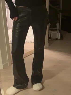Svarta skinnbyxor med raka ben - Säljer ett par svarta skinnbyxor med raka ben och hög midja. Byxorna har en stilren look med fickor bak och stängs med dragkedja och knapp. Perfekta för dig som vill ha en edgy och trendig stil. Har några små slitningar i tyget.