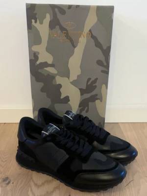  Valentino Garavani rockrunners  - Säljer ett par riktigt schysta svarta sneakers från Valentino Garavani med en mix av mocka och läderdetaljer. Skorna har kamouflagemönster på ovandelen, svarta skosnören och en chunky sula med nitar på hälen. Perfekta för dig som gillar exklusiv streetstyle. Dom är sparsamt använda och är i topp skick. Sko lådan och crease skydd tillkommer. Priset kan diskuteras. Vid någon som helst fråga hör av er ☺️