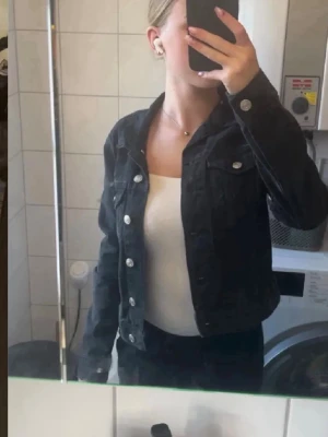 Svart jeansjacka med silvriga knappar - Snygg svart jeansjacka med klassisk krage och silvriga knappar framtill. Jackan har två bröstfickor med lock och knapp samt raka ärmar. Perfekt att slänga över en t-shirt eller linne för en cool look.