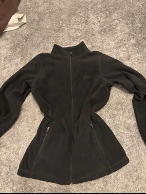 Svart fleecejacka med dragkedja - Mysig svart fleecejacka med hel dragkedja framtill och två fickor med dragkedja. Jackan har hög krage och diskret broderad logga på bröstet. Perfekt för lager-på-lager och håller dig varm under kyliga dagar.