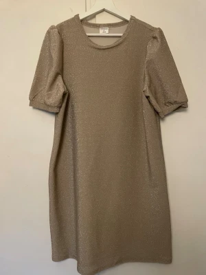 Glittrig beige kortklänning  - Kortklänning i glittrigt beige tyg. Klänningen har puffiga korta ärmar och rund halsringning. Perfekt för dig som vill sticka ut med en enkel men festlig look.