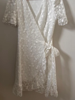 Vit omlottklänning med spets och knyt - Romantisk vit kortklänning i spets med blommigt mönster. Omlottmodell med knyt i midjan, volangkant nedtill och korta ärmar. Perfekt för sommarens alla tillfällen, skolavslutning, student mm.