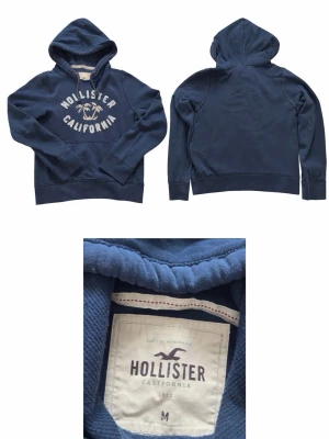Mörkblå hoodie från Hollister - Mörkblå hoodie från Hollister med stor huva och magficka. Framtill står det 'Hollister California' i vitt med broderade bokstäver och palmmotiv. Tillverkad i mjukt bomullsmaterial, perfekt för en avslappnad och cool stil.