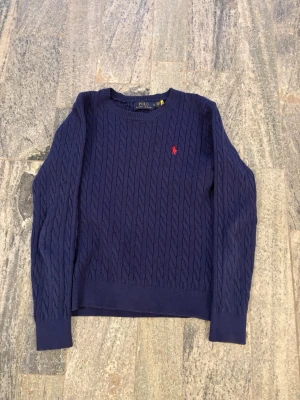 Mörkblå kabelstickad tröja Polo Ralph Lauren - Mörkblå kabelstickad tröja från Polo Ralph Lauren med rund halsringning och röd broderad logga på bröstet. Tröjan har långa ärmar och ribbade muddar vid ärmslut och nederkant. Klassisk och stilren design som passar till mycket.