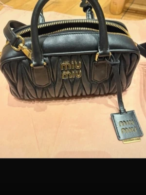 Svart handväska från Miu Miu - Snygg svart handväska från Miu Miu i mjukt läder med quiltad design och guldfärgade detaljer. Väskan har dubbla handtag, dragkedja upptill och en tag med logotyp. Perfekt för dig som gillar lyxiga accessoarer med trendig känsla.