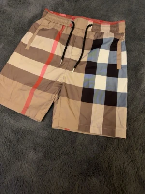 Burberry short  storlek s - Snygga shorts med klassiskt rutigt mönster i beige, brunt, svart, vitt och blått samt en röd detalj. Shortsen har elastisk midja med svart och vit dragsko, sidofickor och en bakficka. Perfekta för en trendig och avslappnad look.