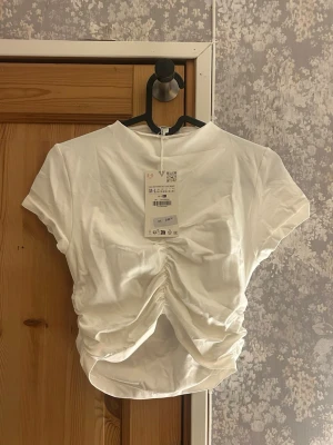 Vit croppad topp med rynkdetalj - Vit kroppad t-shirt med rynkdetalj där framme från Zara. Helt oanvänd med prislapp. Hör av vid frågor!💗