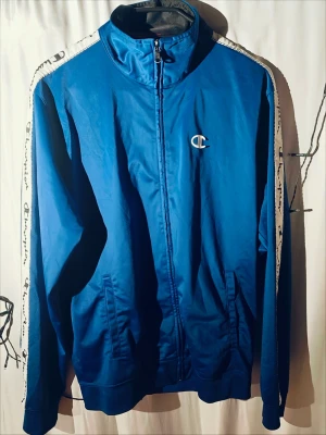 Blå träningsjacka från Champion - Snygg blå träningsjacka från Champion med dragkedja framtill och vita detaljer längs ärmarna där Champion-loggan är tryckt. Jackan har ståkrage, två fickor och ribbade muddar. Perfekt för sport eller chill. Materialet är troligtvis polyester.