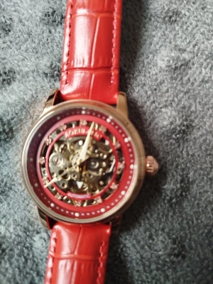 Röd mekanisk klocka med skinnband - Säljer en snygg mekanisk klocka från Olevs med transparent urtavla och röd ram. Klockan har ett rött skinnband med krokodilmönster och guldfärgat boett. Perfekt för dig som gillar detaljer och stil.frakt tillkommer använder mig av Postnord alt hämt.