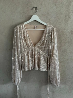 Champagnefärgad paljettopp med v-ringning - Säljer en glittrig champagnefärgad topp med paljetter, djup v-ringning och långa ärmar. Toppen har omlottkänsla och knytband i midjan för extra detalj. Perfekt för dig som vill sticka ut med en festlig look.