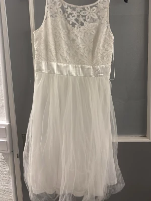 Vit spetsklänning med tyllkjol - Söt vit klänning med blommig spets upptill och en fluffig tyllkjol. Klänningen har ärmlös design och rund halsringning. Perfekt för sommarens alla tillfällen eller studentfirande. Midjan markeras med ett glansigt band.