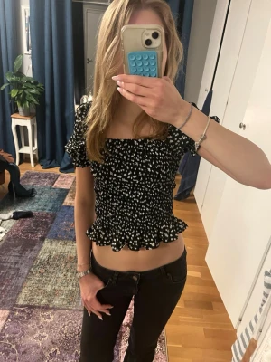 Svart blommig  topp - Söt svart croppad topp med vitt blommönster och volangkant. Toppen har korta puffärmar som kan bäras både på axeln och offshoulder. I storlek 170 men passar mig som vanligtvis har xs 