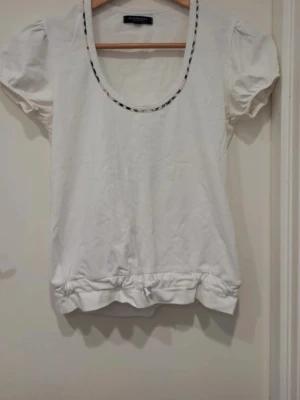 Vit topp från Burberry med puffärm - Snygg vit topp från Burberry med rund halsringning som har klassiskt rutmönstrad kant. Puffärmar och dekorativa detaljer vid fållen ger en söt och trendig look. Tillverkad i mjukt bomullsmaterial, perfekt för en avslappnad men stilsäker outfit.