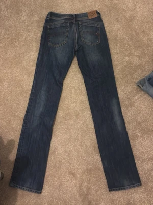 Mörkblå Tommy Hilfiger jeans - Snygga mörkblå jeans från Tommy Hilfiger med raka ben och klassisk femficksmodell. Jeansen har diskreta slitningar och detaljer i brunt läder på bakfickan. Tillverkade i denim med kontrastsömmar och Hilfiger-logga på fickan.