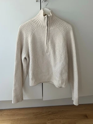 Beige stickad halvzip tröja - Snygg beige stickad tröja med halvzip och hög krage. Tröjan har ett ribbstickat mönster och långa ärmar. Perfekt för lager på lager och ger en clean och stilren look. Nopprig.