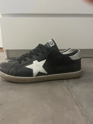 Golden Goose svart/vit sneakers - Svarta sneakers från Golden Goose med vit stjärndetalj på sidan och vit häl. Modellen heter Super-Star och har svarta skosnören samt en låg silhuett. Skorna har en klassisk design med gummisula och är tillverkade i läder.